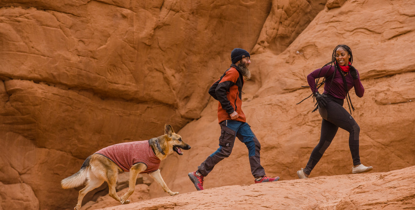 A woman and man run with their dog on a desert trail. 
