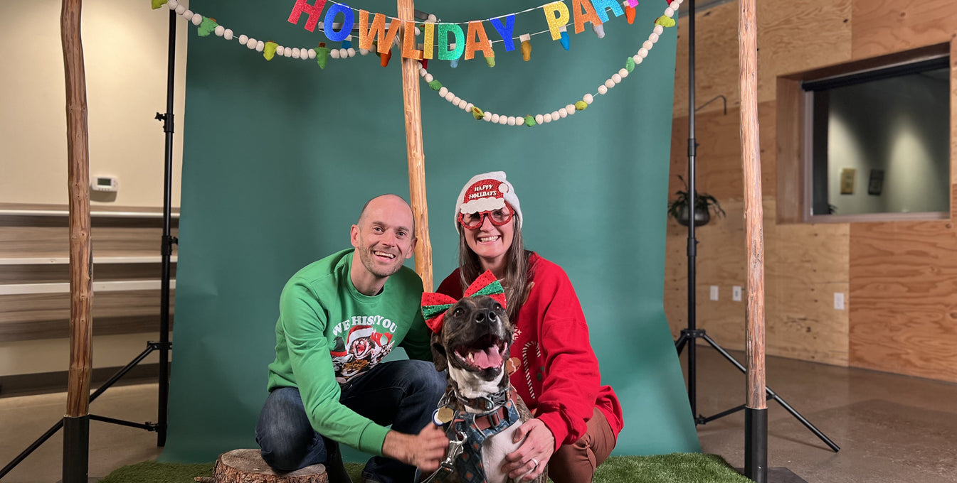 A woman, man, and their dog dressed in holiday outfits pose for a picture at the Ruffwear Howliday Party photo booth. 