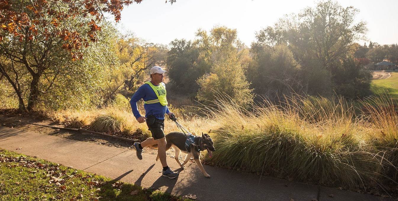 Blind runner and dog in Unifly run along a paved trail with trees.