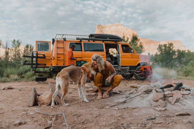 Woman in orange overalls kisses dog in orange front range harness while they sit in front of orange van in desert!
